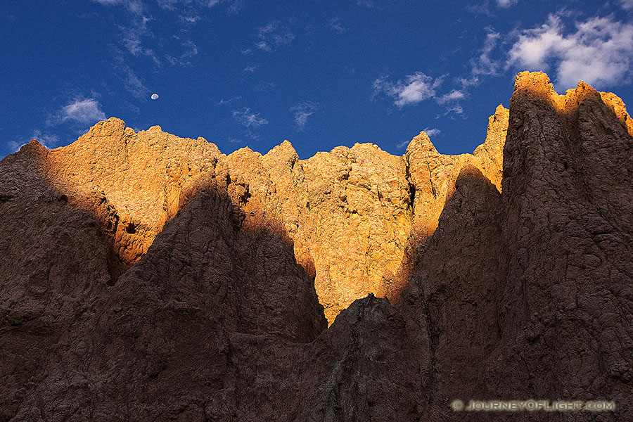 The first warm light of sunrise illuminates the erie rock formations deep in Badlands National Park as a waning moon gentle descends behind. - South Dakota Photography