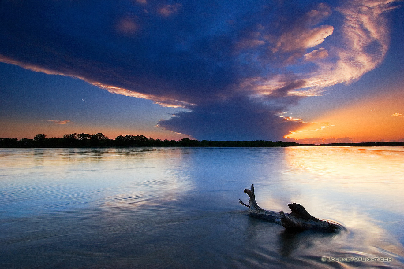 The setting sun shines through a small hole in the clouds as the Platte River flows past a lodged log.  This was taken near Schramm State Recreation Area. - Schramm SRA Picture