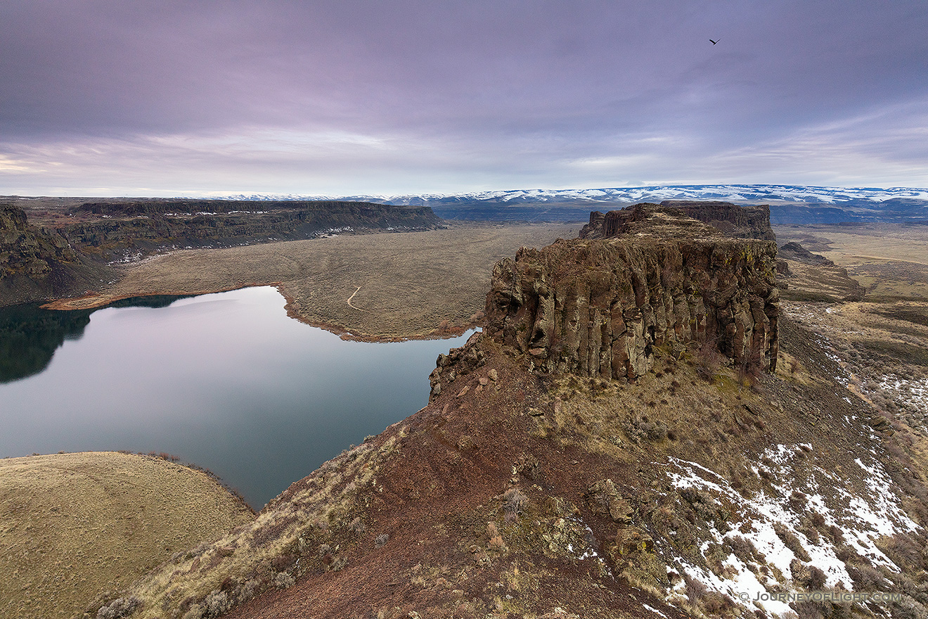 Approximately 17 million years ago lava flowed across what is now eastern Washington into Oregon.  Every few thousand years these lava flows would occur and then stop for long periods of time.  It would deposit a fine grained rock known as basalt.  After the flows stopped permanently large floods ran through this area carving out deep ravines.  Eventually, the Columbia River formed and snaked its way through Central Washington.  This landscape is the Potholes Coulee area in the Quincy Lakes unit of the Columbia Basin Wildlife Area displaying some of the unique landscape caused by these geologic events.  

I captured this image on a February morning.  It was cool, but not cold and a few remnants of a previous snow fall are still visible.  When cresting the cliff, I unfortunately startled a flock of geese in Dusty Lake below.  I took a moment to appreciate the view and then went to work capturing the beauty before me.  In the distance mountains, still covered with snow lay just beyond the unseen Columbia River.  As I was finishing up, a hawk climbed into the sky and then out of sight.  As I packed up I imagined myself as that hawk flying over the unique landscape. - Pacific Northwest Picture